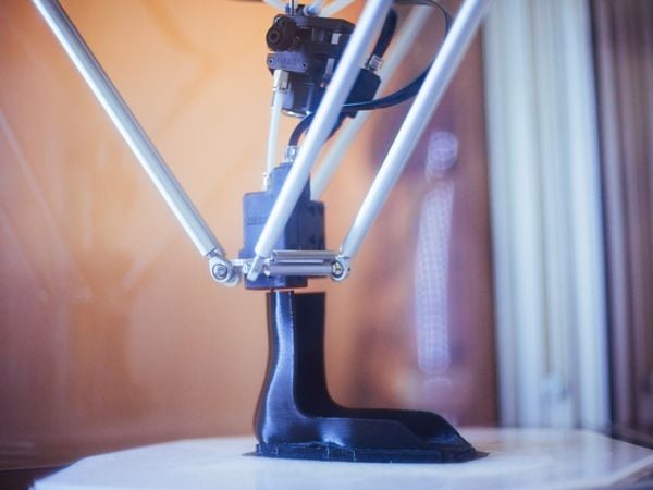 Eine 3D-Orthese wird gedruckt. Sie ist schwarz und steht auf einem weißen Untergrund. Verschiedene Metallteile laufen von oben zusammen und zur Spitze, die die Orthese druckt.